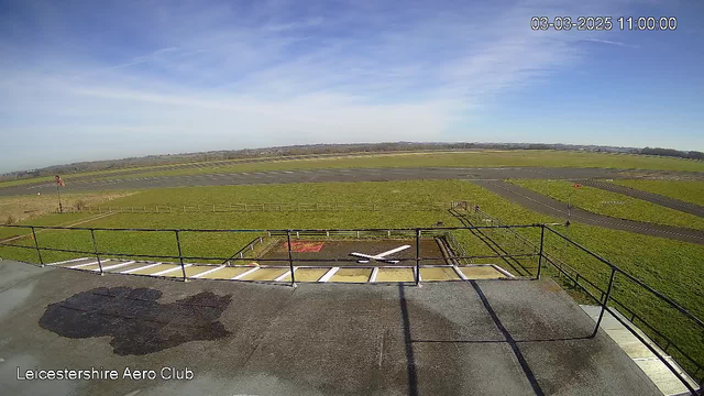 A clear view from a webcam at the Leicestershire Aero Club shows a wide expanse of green grassland and a runway. In the foreground, there is a black surface with white markings resembling an "X." The sky is bright blue with scattered clouds, and a few distant hills are visible on the horizon. There is a windsock to the left, indicating wind direction.