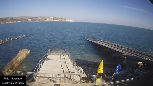 A view of a waterfront scene with calm blue water extending towards the horizon. In the foreground, there is a flat concrete area leading to the water, bordered by a railing. Two small boats, one yellow and one blue, are secured near the shoreline. To the left, a curved wooden pier extends into the water, and in the distance, a rocky coastline with white cliffs is visible under a clear blue sky. The bottom left corner displays a timestamp and location label.