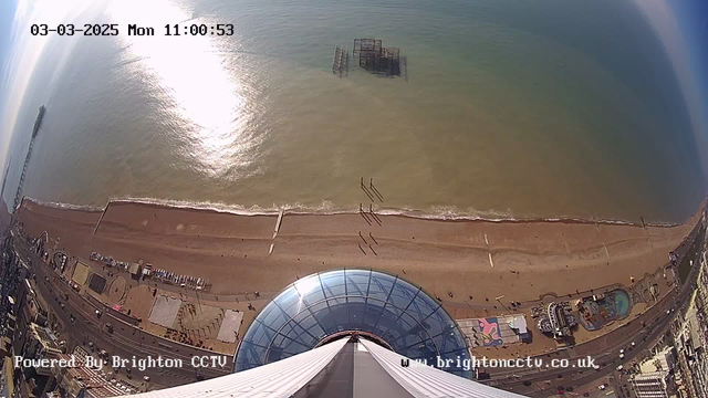 A high-angle view from a webcam overlooking a beach. The ocean is visible with sunlight reflecting on the water's surface. A pier extends into the sea on the left side of the image. Along the sandy beach, there are people walking, and various structures and attractions are visible on the beach front. In the foreground, the top of a circular glass dome is seen. The sky is clear with a hint of blue. The date and time are displayed at the top.