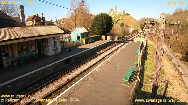 A railway station platform with visible tracks extends into the distance. On the left, there is a stone building with a sloped roof, possibly a ticket office, and a few benches are arranged along the platform. A green shed and hedges are in the background, while a hill with a castle ruin rises prominently in the distance under a clear blue sky. Power lines are visible in the foreground, and the scene is bathed in bright sunlight.