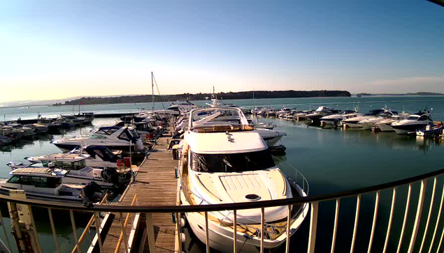 A view of a marina filled with various boats and yachts docked at a wooden pier. The water is calm and reflects the clear blue sky. There are additional boats lined up in the distance, surrounded by hills. The scene is bright and serene, capturing a peaceful moment by the water.