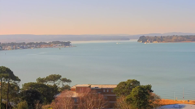 A calm body of water stretches across the image, reflecting a light blue hue under a clear sky. In the distance, a coastal town is visible with several boats docked, and a line of trees borders the waterfront. On the right, a small island features a building and more trees, while the shoreline curves gently towards the horizon, where rolling hills can be seen. In the foreground, there are patches of greenery and a few residential buildings.