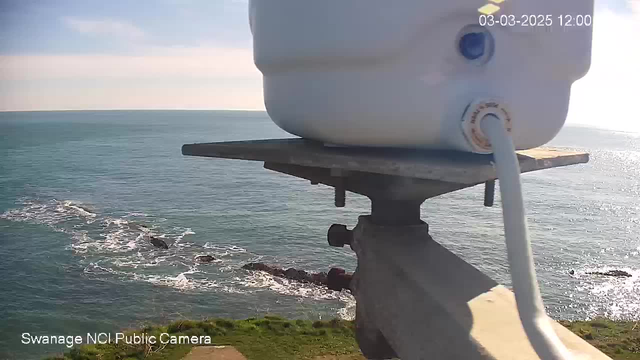 A view of the ocean from a high vantage point, featuring a white container positioned on a metal platform. Below, waves crash against rocky formations along the shoreline. The sky is bright with some clouds, and the water reflects sunlight, creating a shimmering effect. The lower part of the image includes green grass leading up to the rocks. Text at the bottom indicates it's a public camera feed from Swanage.