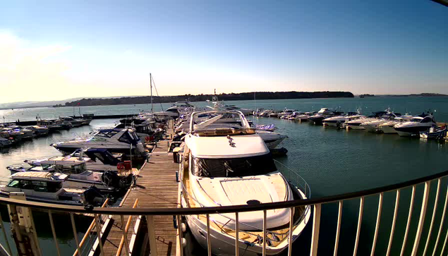 A marina scene with several boats docked along a wooden pier. In the foreground, a large white yacht is visible. Surrounding boats of various sizes and colors are moored along the water's edge. The background features calm blue water and a clear sky. Some land is visible in the distance, suggesting it is a tranquil coastal area.