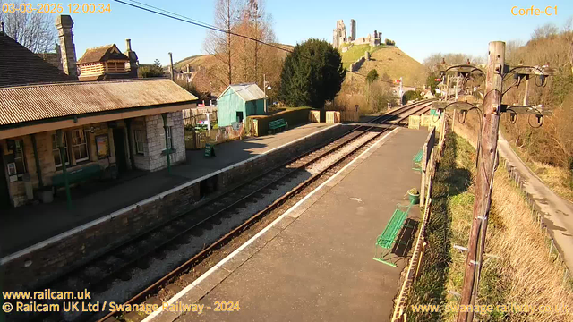 A view of Corfe Castle station on a clear day. The foreground features a platform with a few green benches along the edge. To the left, there's a stone building with a peaked roof, likely a station building, and a small green shed. The railway tracks extend into the distance, with a wooden telephone pole and some trees visible on the right side. In the background, the hill rises to reveal the ruins of Corfe Castle atop it, surrounded by greenery and a clear blue sky.