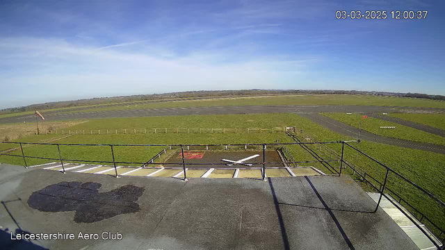 A wide view of an airport runway taken from a high vantage point. In the foreground, there is a viewing platform with a flat surface. To the left, a windsock is visible, indicating wind direction, and an adjacent area features a large, white "X" marking on the ground. The runway extends into the distance, lined with green grass on either side. Blue sky with some scattered clouds is above, and a few distant hills are visible on the horizon. The scene captures a bright, clear day.