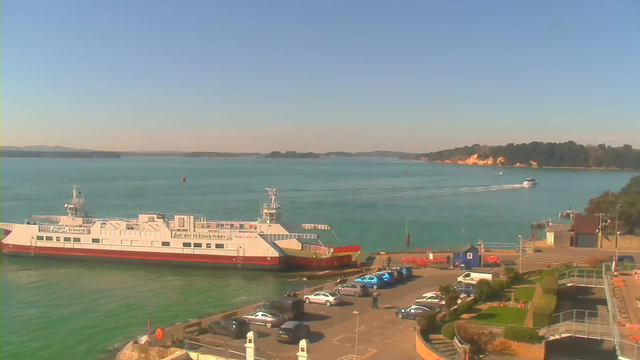 A ferry is docked at a terminal on a sunny day, with calm turquoise water surrounding it. Small boats are seen traveling in the distance. A parking lot with several cars is situated near the ferry terminal, and there are some people walking nearby. In the background, there are green trees and sandy cliffs. The sky is clear and blue, indicating pleasant weather.