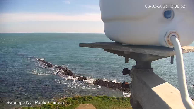 A view of the ocean from a high vantage point, with rocky outcrops jutting into the water. The sea is calm and a vibrant blue, reflecting a clear sky with a few wispy clouds. In the foreground, part of a white cylindrical object is visible, secured to a metal stand. The scene suggests a coastal area, characterized by greenery at the bottom where the land meets the water. The image is timestamped, indicating the date and time.