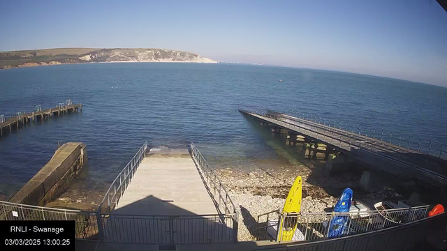 A view of a coastal area featuring calm blue water and a light blue sky. In the foreground, there are two wooden piers extending into the water, one of which has a gentle slope leading down to the shore. There are some kayaks in bright yellow, blue, and orange, resting on a small beach made of pebbles and rocks. The background shows a distant coastline with cliffs and grassy hills. The scene is clear and sunny, indicating a pleasant day.