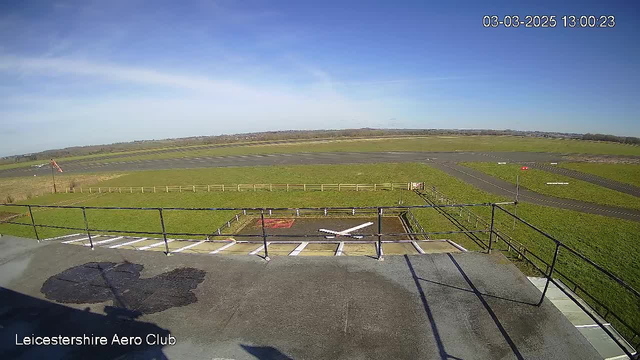 A wide view of a grassy airfield under a clear blue sky. In the foreground, there is a rooftop structure with railings, and a helicopter landing pad marked with a white cross. The horizon features a few trees and further away, there are runways for aircraft. A small windsock is visible, indicating wind direction. The overall scene conveys a bright, sunny day at Leicester Aero Club.