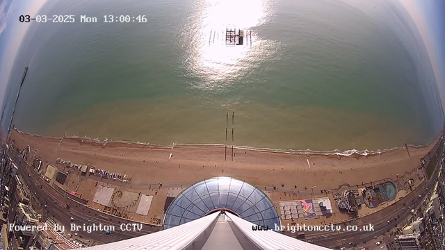 A view from above looking down at a beach with golden sand and gentle waves, a pier with wooden structures extending into the water, and various buildings and attractions along the shoreline. The sun reflects off the ocean surface, creating a shimmering effect. The sky is partially clear, and there are a few clouds visible. The date and time are displayed at the top of the image.