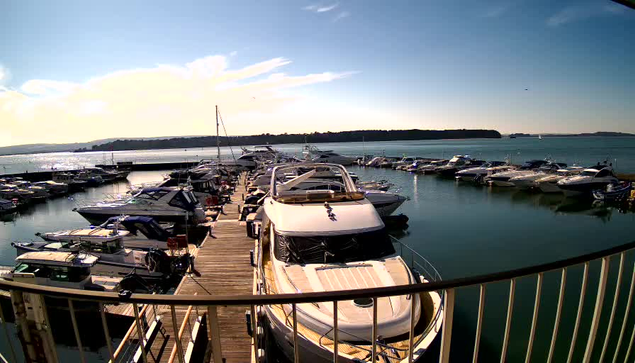 A marina scene shows numerous boats docked in calm water under a clear blue sky with wispy clouds. In the foreground, a white yacht is prominently placed, with several other boats of various sizes and colors moored nearby. The dock extends along the left side, leading further into the marina where more boats are visible. The background features a distant landmass and a few boats sailing on the water.