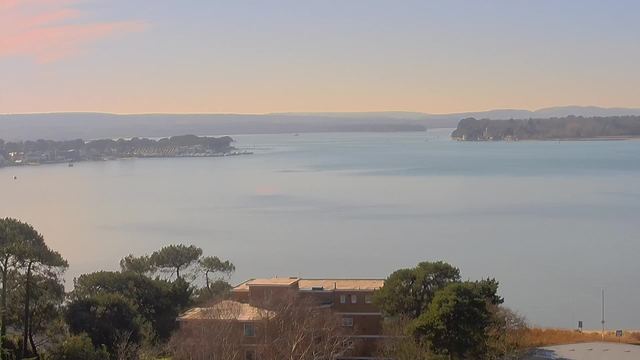 A calm body of water reflects soft pastel colors of a sunrise or sunset, with a shoreline lined by trees. In the distance, a marina with boats is visible, alongside houses and docks. The horizon features gentle hills or mountains against a clear sky. The foreground includes green trees and a low building made of light-colored material. The overall scene conveys tranquility and natural beauty.