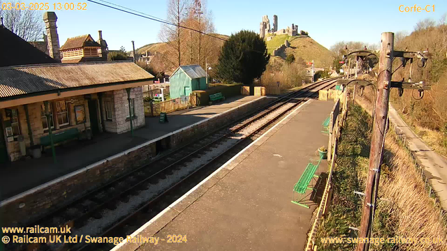 A railway station platform is depicted in the image, with a background of rolling hills and ruins. The platform holds several green benches, and a small building with a sign is visible, labeled “GIRL.” To the right, there's an assortment of trees and a blue shed. The railway tracks run along the left side of the image, and the scene is bathed in bright sunlight, indicating clear weather. In the distance, the remains of a castle are perched on a hill, adding a historical element to the landscape.