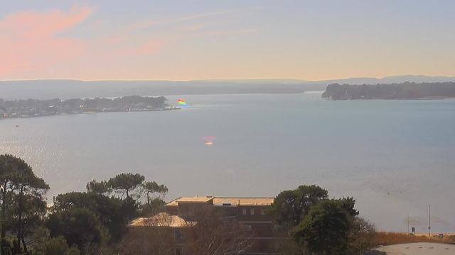 A scenic view of a calm body of water under a clear sky, reflecting soft colors of pink and blue. In the foreground, there are low trees with green foliage, and an outline of a building with a tan roof. In the distance, a small harbor is visible with a few boats and a colorful rainbow reflection on the water's surface. The horizon features gently rolling hills.