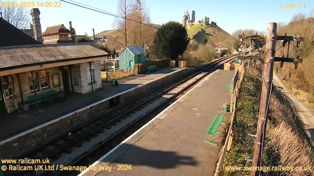 A view of Corfe Castle from a railway station. The scene features a platform with two green benches and a small wooden shed. In the background, the castle ruins are visible atop a green hill. The sky is clear and blue, and a set of railway tracks runs along the platform. There are trees and bushes surrounding the area, providing a natural backdrop.