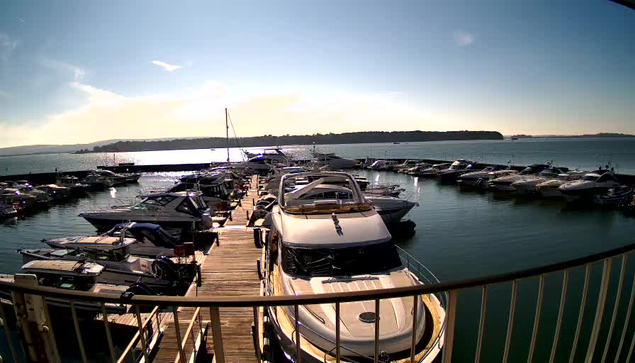 A marina scene featuring numerous boats docked in calm water. A wooden pier leads into the marina, with several boats lined up on either side. The sky is clear with some distant hills visible in the background, and the sunlight reflects off the water, creating a shimmering effect.