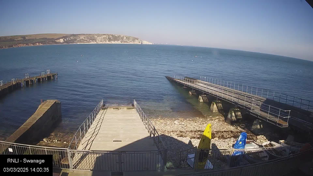 A clear view of a coastal area featuring a calm sea under a blue sky. In the foreground, there is a wooden pier with a ramp leading down to the water, surrounded by a rocky shoreline. Several boats are visible on the beach, including a yellow kayak and a blue motorboat. The water appears tranquil, with a few buoys floating. Cliffs are visible in the background, and the entire scene is illuminated by sunlight.