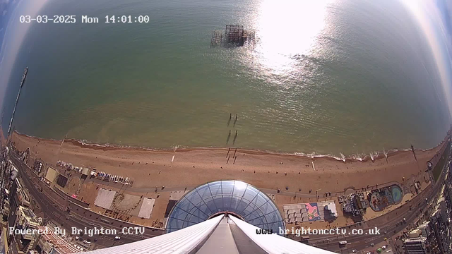 A high-angle view from a webcam showing a beach and the sea. The sandy beach stretches across the lower part of the image with several people walking, and some small structures near the water's edge. An amusement pier is visible in the distance. The sea is calm with gentle waves, while the sun reflects off the water, creating a shimmering effect. Above the beach, there is a clear blue sky with a few clouds. The upper part of the image includes a circular glass structure that appears to be part of a building or observation deck. The time and date are displayed in the top left corner.