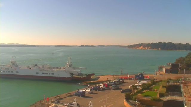 A clear view of a harbor scene on a sunny day. In the foreground, there is a ferry docked at a terminal, surrounded by calm turquoise water. To the right, several cars are parked in a lot, and there are green landscaped areas with palm trees. In the background, a line of hills and trees can be seen along the coastline, with a light sandy shore visible. The sky is bright blue with minimal clouds, suggesting a pleasant day.