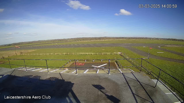 A view from a high vantage point overlooking an airfield. The foreground includes a fenced area, marked with a white cross and some red ground markings. Beyond this, there is a wide expanse of green grass leading to a runway and parking areas in the distance, bordered by wooden fences. The sky is bright blue with a few white clouds scattered throughout. A flagpole is visible to the left, with the flag fluttering in the wind. The time and date displayed on the image indicate it is 2:00 PM on March 3, 2025.