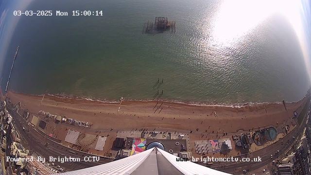 Aerial view of a beach scene on a sunny day. The camera is positioned high above, looking straight down at the sandy shore. The beach is populated with people walking along the water's edge. A pier extends into the water in the background, with some structures visible on the sand below. The sea appears calm and glistens in the sunlight. Colorful structures and rides are visible along the shoreline, indicating amusement attractions. The image is timestamped at 15:00 on March 3, 2025.