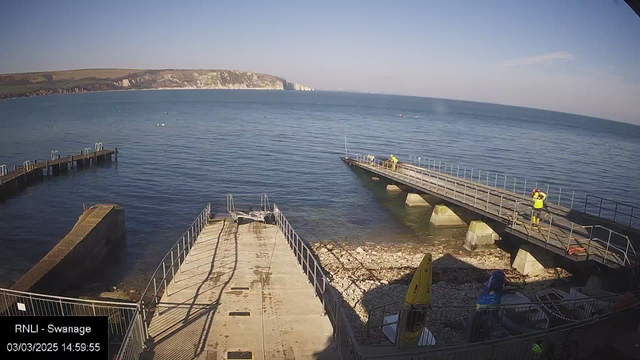 A view of the sea from a coastal area, featuring a clear blue sky and gentle waves. In the foreground, there are two piers extending into the water, one with a ramp and the other with railings. A few people, dressed in yellow safety gear, are visible on the right pier, working. The shore is rocky with some small boats and equipment along the edge. The cliffs are visible in the background, adding natural scenery to the landscape.