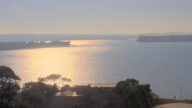 A serene body of water reflecting sunlight, creating a shimmering, golden appearance. In the foreground, there are silhouettes of trees and structures. The horizon features a distant shore with gentle hills, and there are small boats in the water. The sky is mostly clear with a soft gradient, transitioning from light blue to pale hues near the horizon.