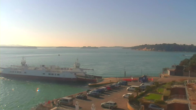 A calm harbor scene featuring a large ferry docked at a pier. The water is a light blue with gentle ripples. In the foreground, there is a parking lot filled with various vehicles, mostly cars, with a few near the water's edge. In the background, distant hills and trees can be seen along the shoreline under a clear sky, suggesting a warm, sunny day.