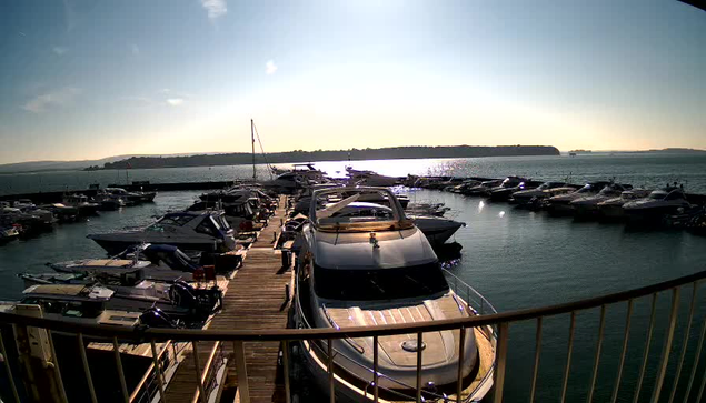 A view of a marina filled with boats, situated on a calm body of water. Several yachts and smaller boats are docked along a wooden pier, extending into the water. The sunlight reflects off the water's surface, creating a shimmering effect. In the background, rolling hills can be seen, partially silhouetted against a clear sky.