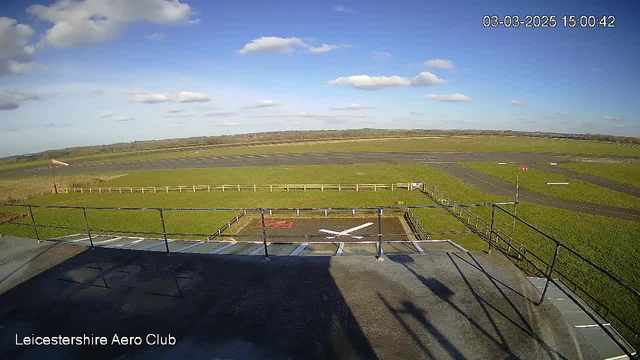 A wide view from a webcam at Leicestershire Aero Club shows a grassy area surrounding a runway. In the foreground, there is a helipad marked with a large red "H" and white cross. A rail fence borders the grassy area, and a windsock is visible near the runway. The sky is clear with scattered white clouds, and the landscape stretches into the distance with fields and a faint outline of trees. The time stamp reads March 3, 2025, at 3:00 PM.