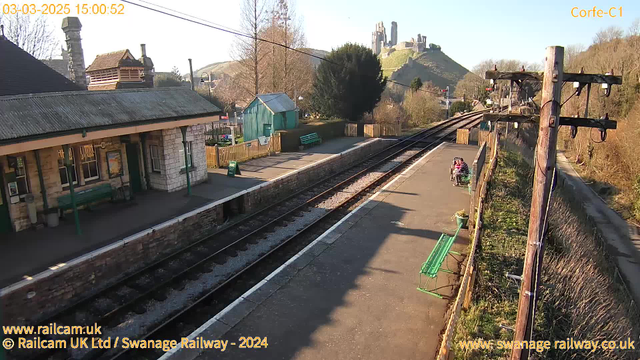 A train station platform is shown with a stone building on the left featuring a sloped roof and large windows. There is a green wooden bench along the platform and a sign indicating an exit. On the platform, two people are seated, and a train track runs alongside the platform with gravel. In the background, a hill rises up with a castle ruin at the top. The sky is clear and bright, indicating daylight.