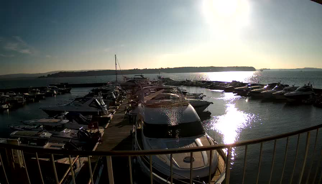 A panoramic view of a marina on a clear, sunny day. Several boats and yachts are moored in calm water, with the sun reflecting off the surface. In the background, gentle hills can be seen on the horizon. The scene conveys a tranquil and vibrant atmosphere.