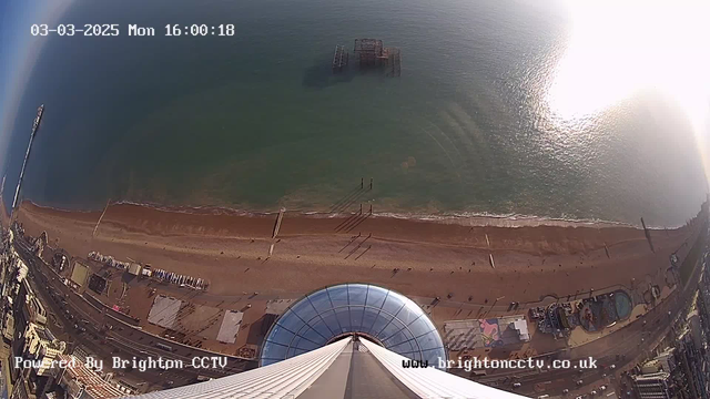 A high-angle view of a sandy beach and the sea. The image shows a curved glass structure in the foreground, likely part of an observation tower. Below, the beach is lined with people, and further out in the water, remnants of a pier structure can be seen. The sea appears calm, with gentle waves reflecting sunlight. In the distance, a boardwalk with various structures is visible. The time and date are displayed at the top of the image.