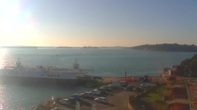 A scenic view of a harbor during daytime. In the foreground, several parked cars are visible along a paved area. To the left, a large white ferry boat is docked. The water is calm with gentle ripples reflecting sunlight, while a distant shoreline and hills can be seen in the background. The sky is clear with a soft gradient from blue to light yellow near the horizon.