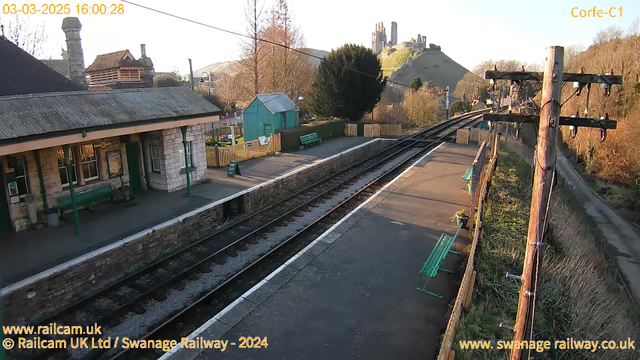 A view of a railway station platform in Corfe, UK, captured by a webcam in the afternoon light. The station features a stone building with a sloped roof, a green bench, and a poster on the wall. On the platform, there are several green benches and a sign that says "WAY OUT." In the background, a hillside is visible, with a castle ruins perched atop it, surrounded by trees and shrubs. The scene is mostly clear, showcasing the tracks leading off into the distance.