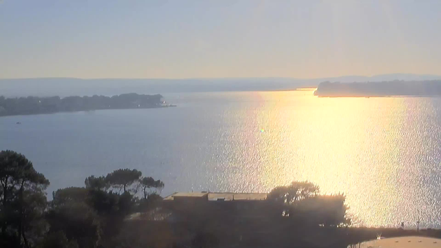 A serene view of a lake or sea under soft sunlight, with the water reflecting glimmers of light. The horizon features distant hills, while trees line the edge of the water, creating a tranquil atmosphere. In the foreground, there is a faint outline of a building or structure, blending with the natural surroundings. The overall scene conveys calmness and natural beauty.