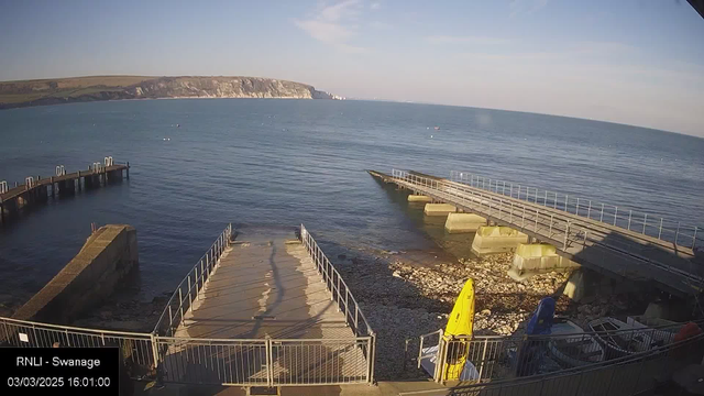 A view of a waterfront scene at Swanage, with calm blue water extending to the horizon. In the foreground, there are two ramps leading to the water, one made of concrete and the other with a flat surface. A small yellow kayak is positioned to the right, while several boats are lined up in a nearby area. The backdrop features a rocky coastline and grassy hills under a clear sky. The timestamp displays the date and time as March 3, 2025, at 16:01.