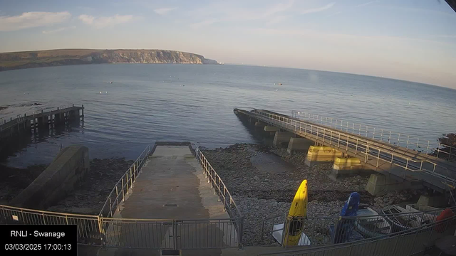 A view of a calm sea and shoreline captured from a webcam. In the foreground, there is a stony area with a ramp leading down to the water. To the left, a small jetty extends into the water, while several kayaks are stored on the right side, with one bright yellow kayak prominently displayed. In the background, a hilly coastline can be seen, under a mostly clear sky with a few clouds.