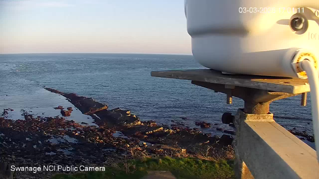 A panoramic view of the coastline featuring rocky outcrops extending into tranquil blue waters. In the foreground, a white webcam mounted on a gray platform is visible. The setting sun casts a soft light across the scene, with gentle waves lapping at the shore. The land in the foreground is grassy, leading up to the rocky area. The sky is mostly clear, transitioning from light blue to a warmer hue near the horizon.