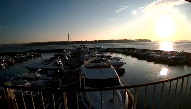 A marina scene at sunset, with numerous boats docked in the water. The sun is setting on the horizon, casting a golden light on the surface of the water. Silhouettes of hills and trees are visible in the background, while a few clouds are scattered in the sky. The foreground features various boats lined up on the dock, creating a sense of tranquility in the evening atmosphere.