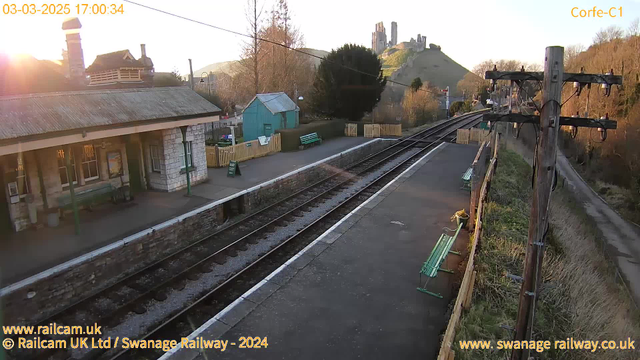 A view of a railway station with a rustic stone building on the left, featuring a sloped roof and green wooden benches. The platform is lined with tracks that extend into the distance. Opposite the station, there are several green benches along a path. In the background, a hill rises with jagged castle ruins visible at the summit, lit by the warm glow of the setting sun. The scene is framed by trees and fences, creating a tranquil landscape.