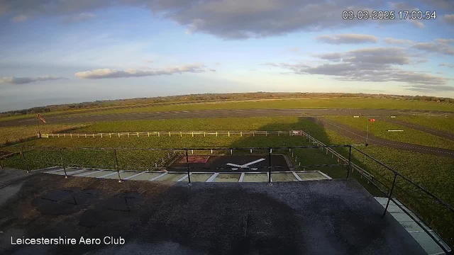 A wide view of an aerodrome captured from above. In the foreground, a helipad marked with a white "X" is visible on a gray surface. There are green fields surrounding the helipad and a white rail fence. In the background, a runway stretches across the image with a small red object situated on the side. The sky is partly cloudy, with patches of blue and a few white clouds. The timestamp in the corner shows the date and time as March 3, 2025, at 5:00 PM.