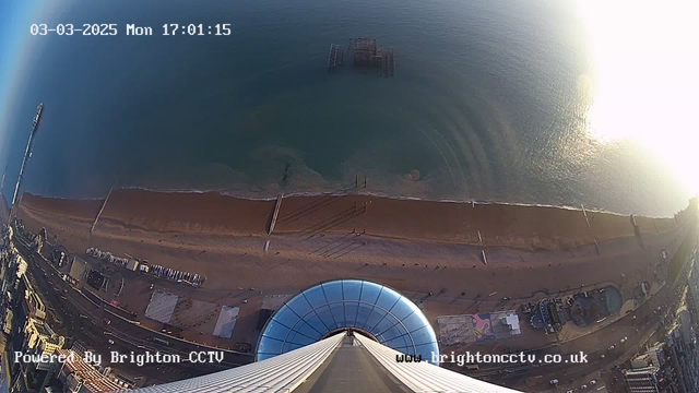 A view from a high point looking downwards towards the beach and ocean. The image shows a circular glass structure at the bottom, which appears to be part of a tower or attraction. The sandy beach extends along the bottom, with some scattered people. In the distance, the ocean is visible, with gentle waves and a pier partially submerged in water. Various beachside buildings and attractions are seen along the shoreline. The sky is clear with a few wispy clouds, and the time stamp indicates it is late afternoon.