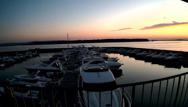 A tranquil marina at sunset, featuring numerous boats moored at a dock. The water is calm, reflecting the soft hues of an orange and pink sky. In the background, grassy hills fade into the distance, while a pier extends into the water. The scene captures a peaceful atmosphere as daylight transitions to dusk.