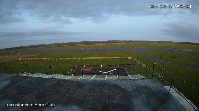 A view from a webcam at Leicestershire Aero Club showing a large, open grassy area with a runway in the distance. In the foreground, there is a black surface with a white cross indicating a helipad. Nearby, a wooden fence marks the boundary of the aerodrome. The sky is partly cloudy, and it appears to be late afternoon with soft lighting. The grass is lush and green, and there are no visible aircraft in the area.