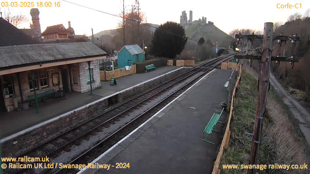 A view of Corfe Castle railway station, featuring a platform with several green benches and a stone wall. There are railway tracks running through the center of the image, leading into the distance. To the right, a wooden pole with wires and lights stands next to the tracks. In the background, Corfe Castle is visible on a hill, surrounded by trees and a sunset sky. The image displays a serene, rural setting, capturing the essence of the station at dusk.