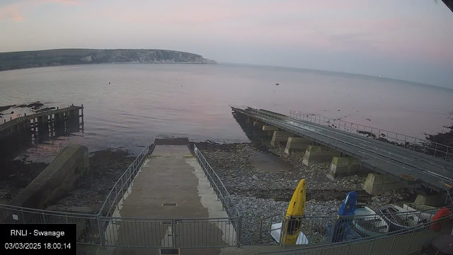 A coastal scene showing calm water under a pastel sky during sunset. In the foreground, there is a ramp leading down to the shore, flanked by rocky areas and a small dock. On the right side, two boats are visible, one yellow and one blue, parked on a stone surface. In the distance, a cliff rises on the horizon, with the sea gently lapping at the shore. The date and time indicate it is March 3, 2025, at 18:00:14.