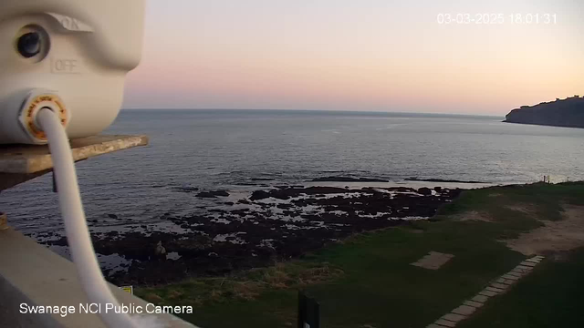 The image shows a coastal landscape at sunset. In the foreground, there is a white camera mounted on a silver platform, with a cable extending downwards. The background features a calm sea reflecting the soft colors of the sky, which transitions from light pink to blue. Scattered rocks are visible in the water, and a patch of green grass borders the shore. To the right, a cliff rises above the water. The bottom left corner of the image includes the text "Swanage NCI Public Camera." The date and time are displayed at the top right corner.