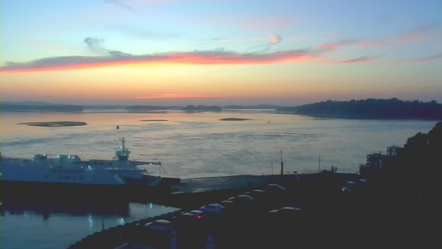 A tranquil scene at dawn showing a body of water reflecting soft hues of pink, orange, and blue in the sky. A large ferry is docked near the left side of the image, with a few lights still glowing on its deck. In the background, scattered islands are visible on the water, and silhouettes of distant hills can be seen under the colorful sky. The foreground includes a dark outline of vehicles parked along the waterfront, framing the peaceful landscape.